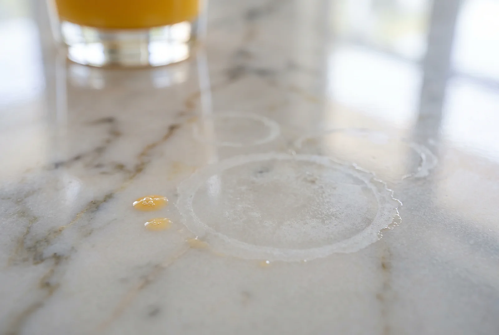 Dull etch rings left on a polished marble countertop by a glass of orange juice, with the acid still beading beside them