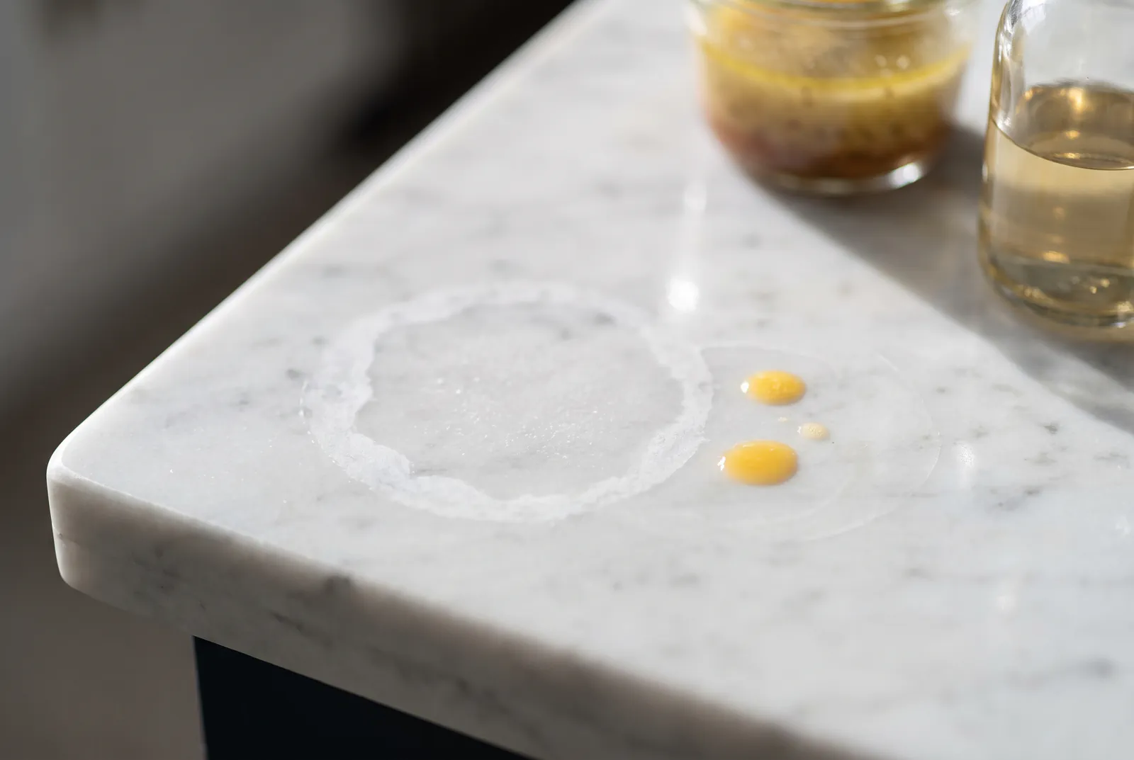 Professional stone sealing: a dull etch ring on a white Carrara marble countertop, with oil droplets and a vinaigrette jar beside it