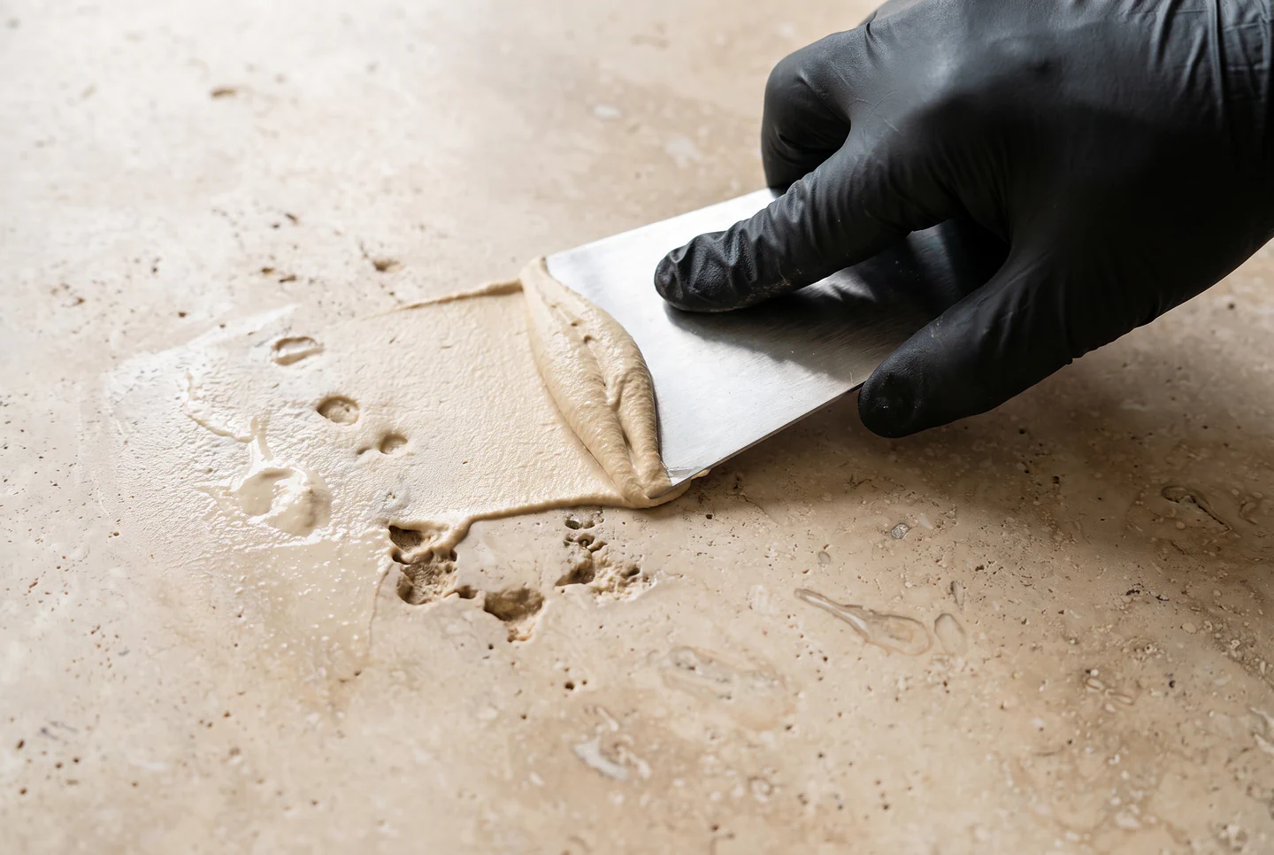 Gloved hand pressing color-matched fill into open pits on a travertine floor in Tampa